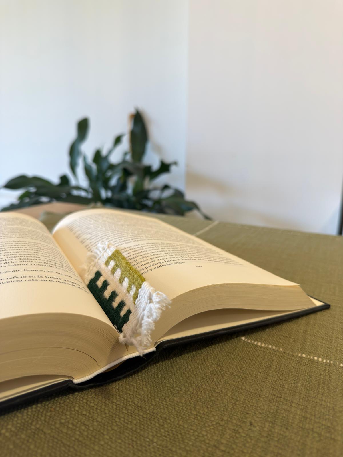 Open book with a green bookmark on a green surface, with a plant in the background.