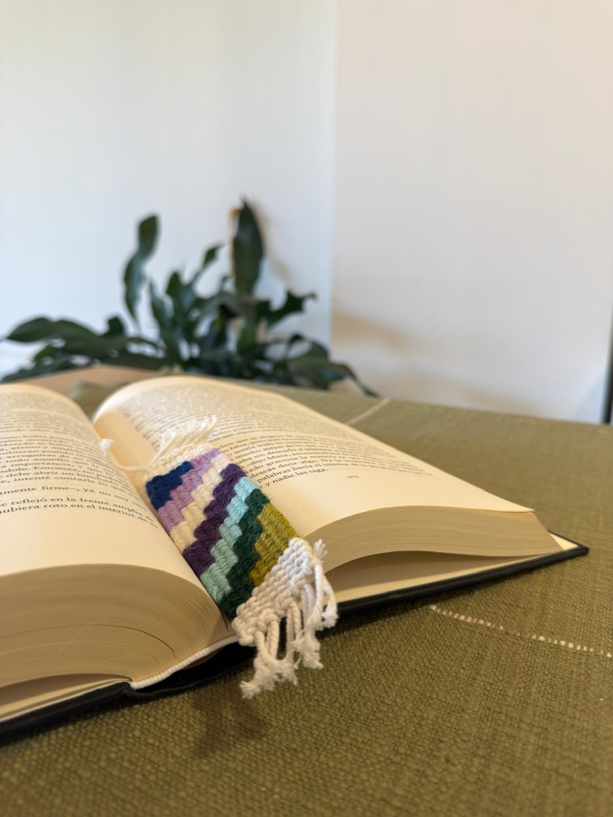 Open book with a colorful knitted bookmark on a green surface, blurred plant in the background