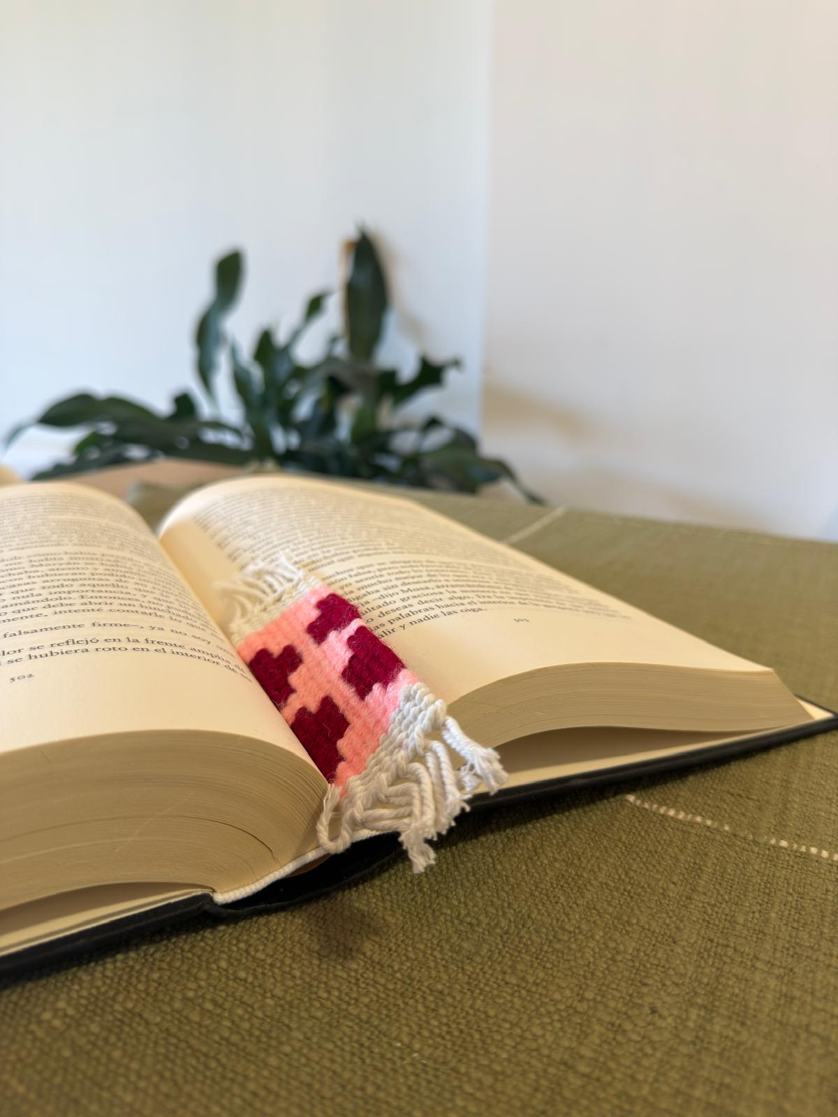 Open book with a colorful woven bookmark on a green surface, with a plant in the background.