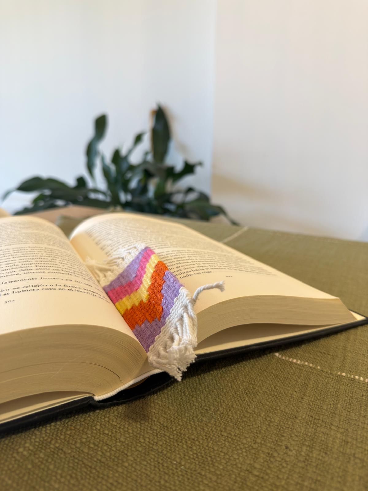 Open book with a colorful knitted bookmark on a green surface, plant in the background