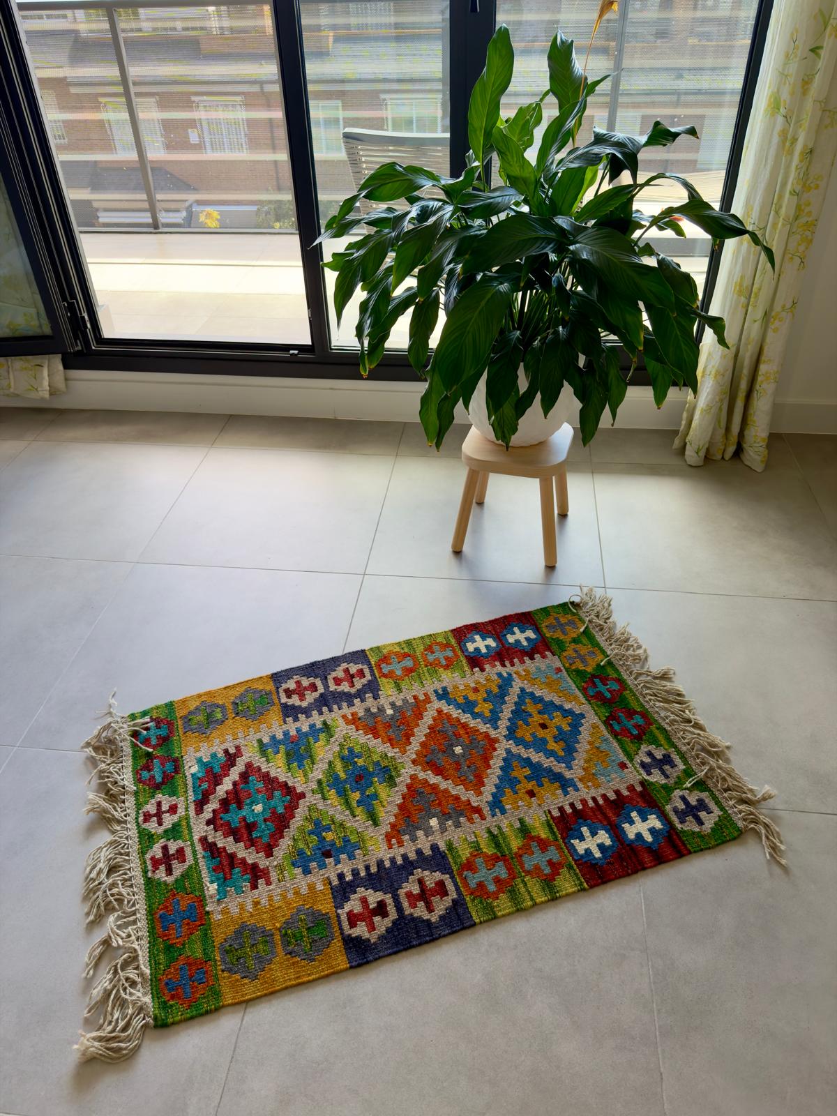 Colorful geometric-patterned rug on a tiled floor with a plant and stool in the background.
