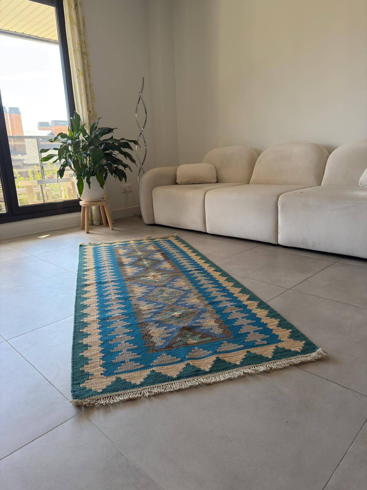Blue and beige patterned rug on a tiled floor with a white sofa and plant in the background.