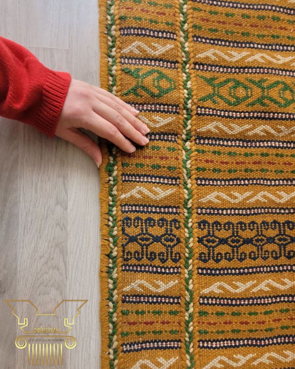 Patterned rug being held by a person on a wooden floor, with a brand logo in the corner.